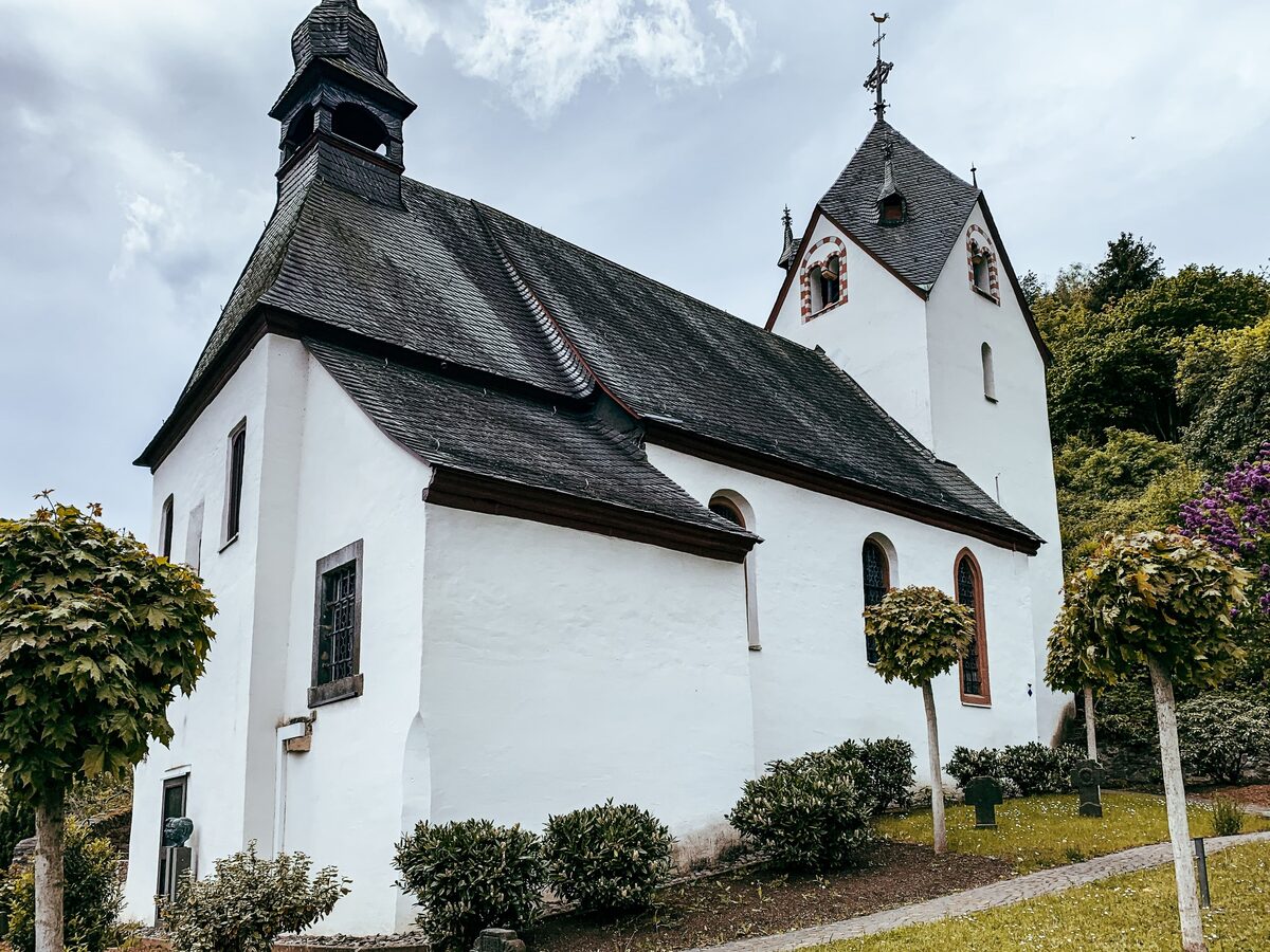 Alte katholische Pfarrkirche St. Bartholomäus in Sankt Aldegund, seitlich fotografiert vor hellem Himmel.